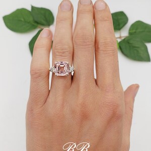 Puede incluir: Anillo con una gran gema rosa de talla cuadrada, flanqueada por dos gemas claras triangulares. El anillo est&aacute; engarzado en una banda plateada. Las iniciales "BB" est&aacute;n en escritura blanca en la parte inferior.