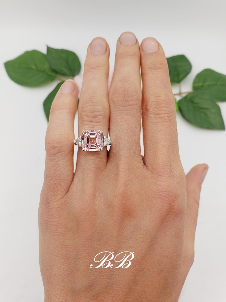 Puede incluir: Anillo con una gran gema rosa de talla cuadrada, flanqueada por dos gemas claras triangulares. El anillo est&aacute; engarzado en una banda plateada. Las iniciales "BB" est&aacute;n en escritura blanca en la parte inferior.