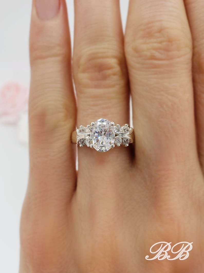 May include: A gold ring with a large oval-shaped center stone and smaller round stones on the band. The ring is on a person's finger.