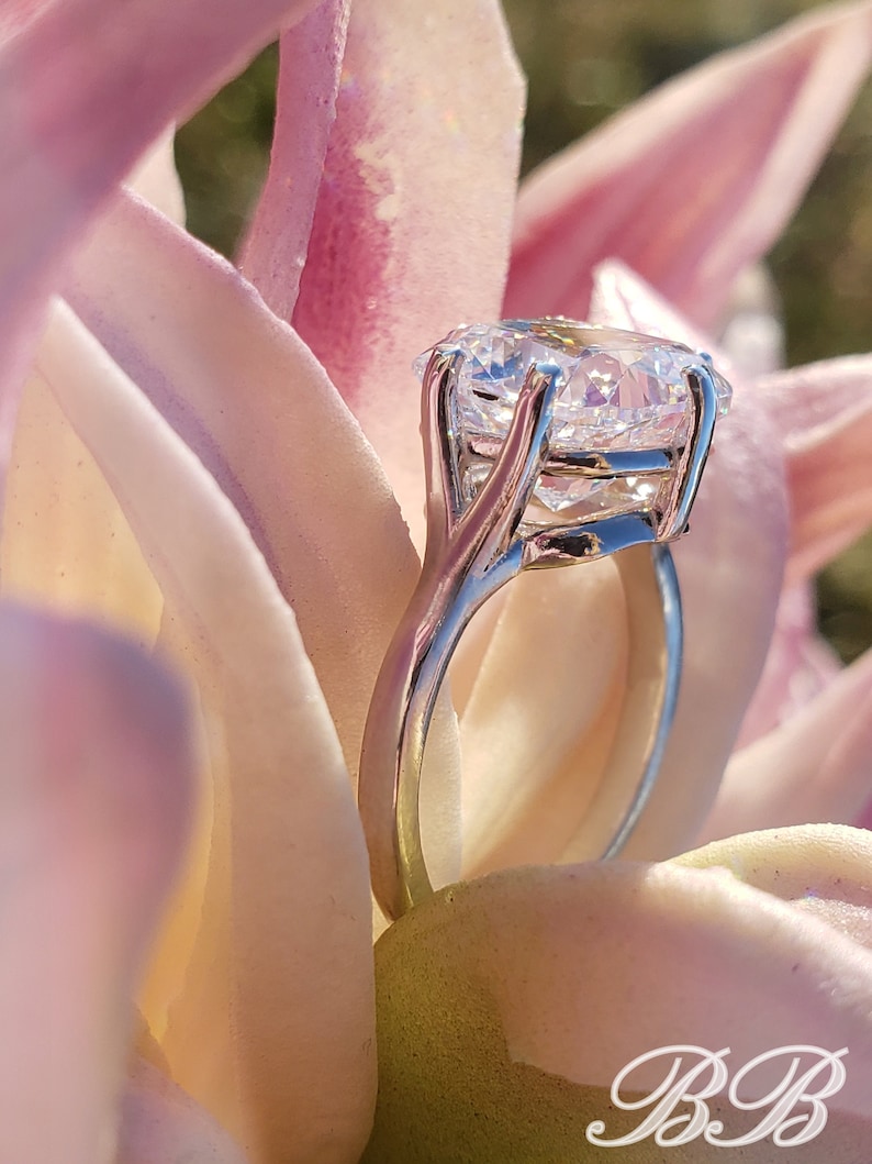 May include: A silver engagement ring with a large, clear, round diamond set in a prong setting. The ring is resting on a pink flower.