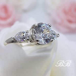 May include: A silver ring with a large round center stone and two smaller pear-shaped stones on either side.