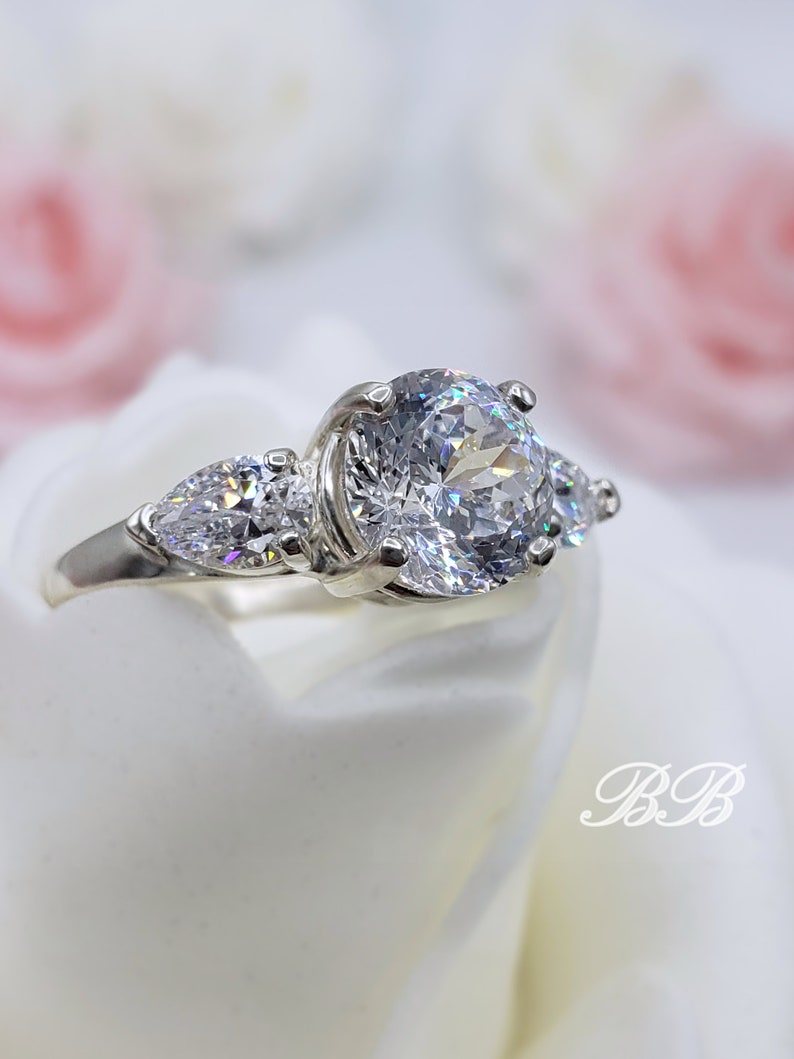 May include: A silver ring with a large round center stone and two smaller pear-shaped stones on either side.