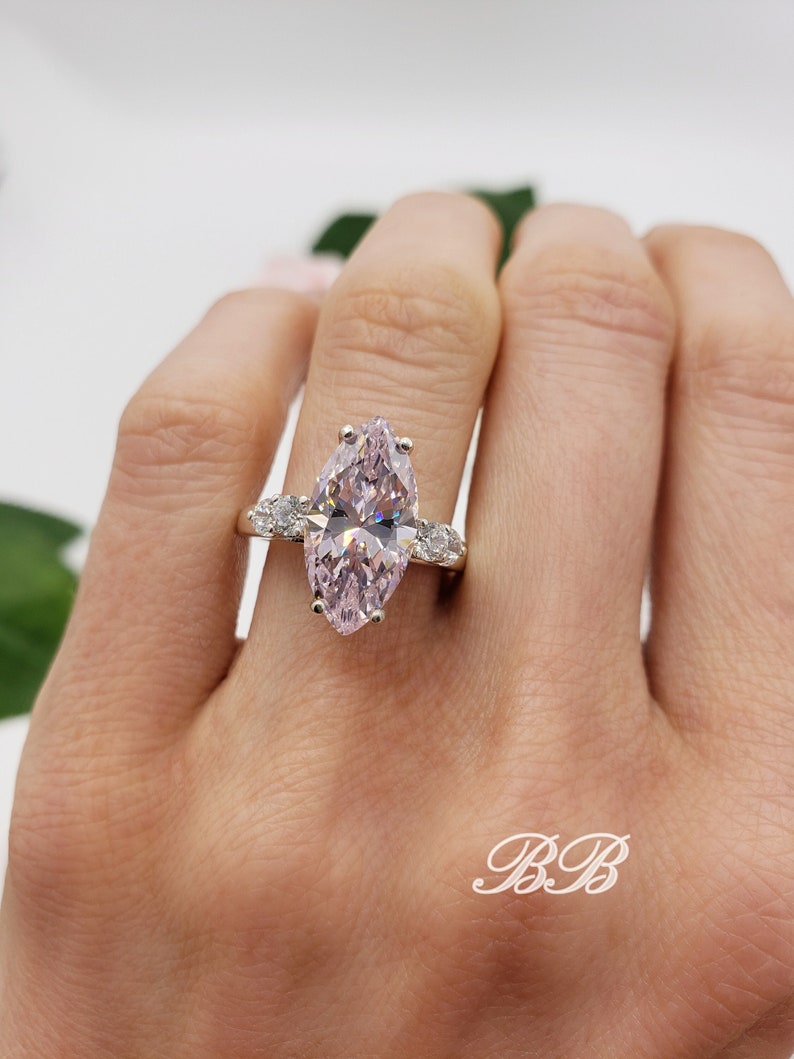 K&ouml;nnte beinhalten: Ein birnenf&ouml;rmiger Verlobungsring mit einem gro&szlig;en rosafarbenen Mittelst&uuml;ck und kleineren klaren Steinen auf der Ringschiene. Der Ring ist in Silber gefasst.