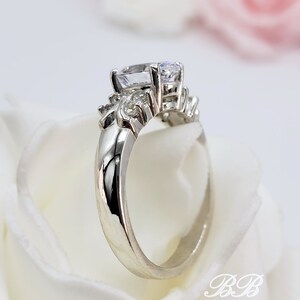 May include: A silver ring with a large, clear, oval-shaped center stone and smaller, clear stones on the band. The ring is set on a white background.