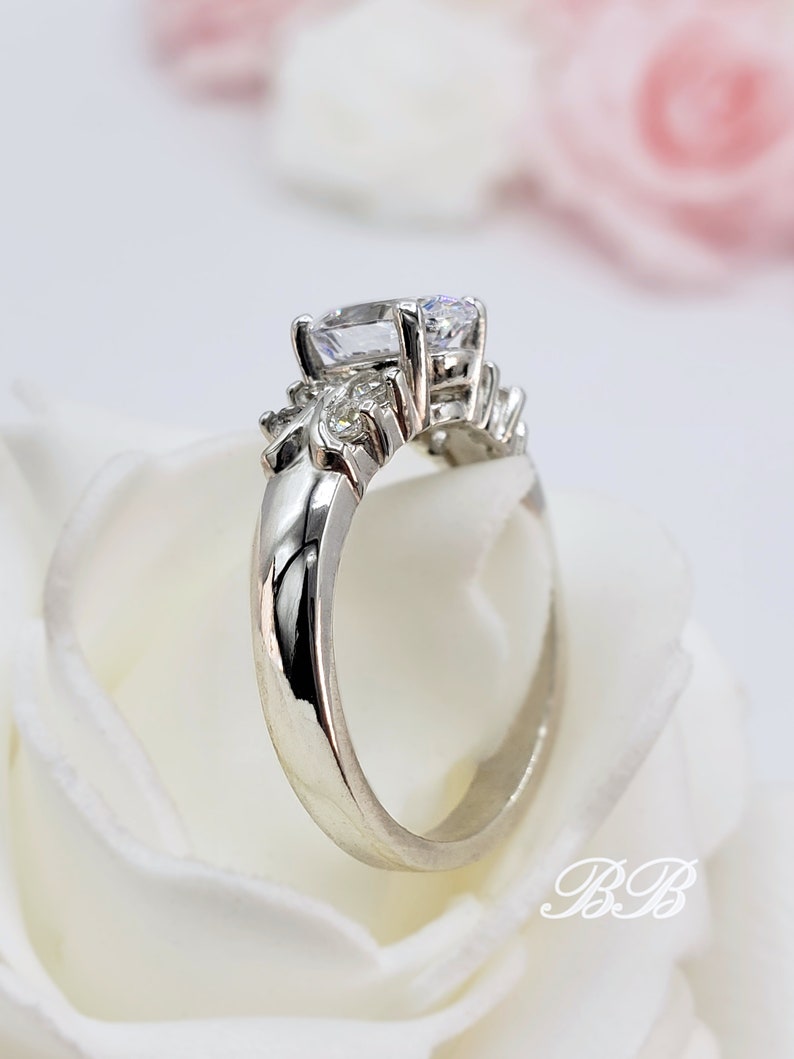 May include: A silver ring with a large, clear, oval-shaped center stone and smaller, clear stones on the band. The ring is set on a white background.