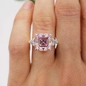 Puede incluir: Un anillo plateado con una gran piedra preciosa rosa claro cuadrada en el centro. Dos piedras preciosas claras triangulares m&aacute;s peque&ntilde;as flanquean la piedra principal. El anillo est&aacute; en un dedo. Las iniciales "BB" est&aacute;n en la parte inferior.