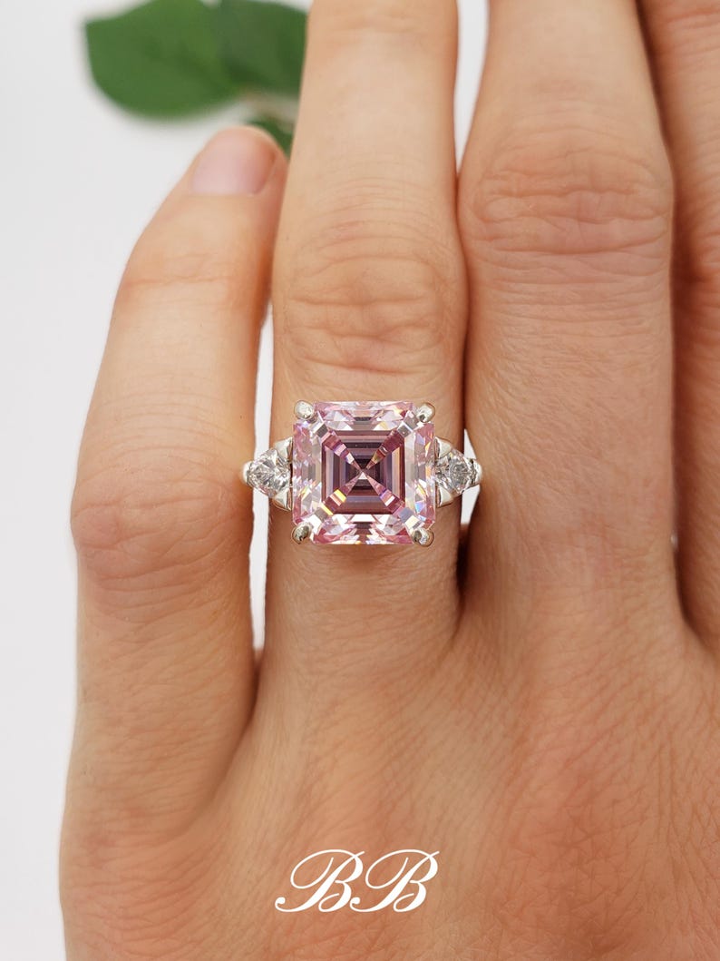 Puede incluir: Un anillo plateado con una gran piedra preciosa rosa claro cuadrada en el centro. Dos piedras preciosas claras triangulares m&aacute;s peque&ntilde;as flanquean la piedra principal. El anillo est&aacute; en un dedo. Las iniciales "BB" est&aacute;n en la parte inferior.