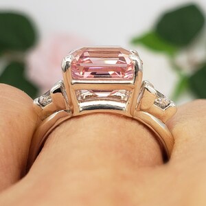 Puede incluir: Primer plano de un anillo con una gran piedra preciosa rectangular de color rosa claro. El anillo tiene una banda plateada y dos piedras preciosas m&aacute;s peque&ntilde;as y claras a cada lado de la piedra principal. Las iniciales "BB" est&aacute;n en la parte inferior.