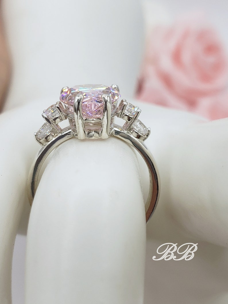 Puede incluir: Un anillo de plata con una gran piedra preciosa rosa de forma ovalada rodeada de piedras preciosas m&aacute;s peque&ntilde;as y transparentes.
