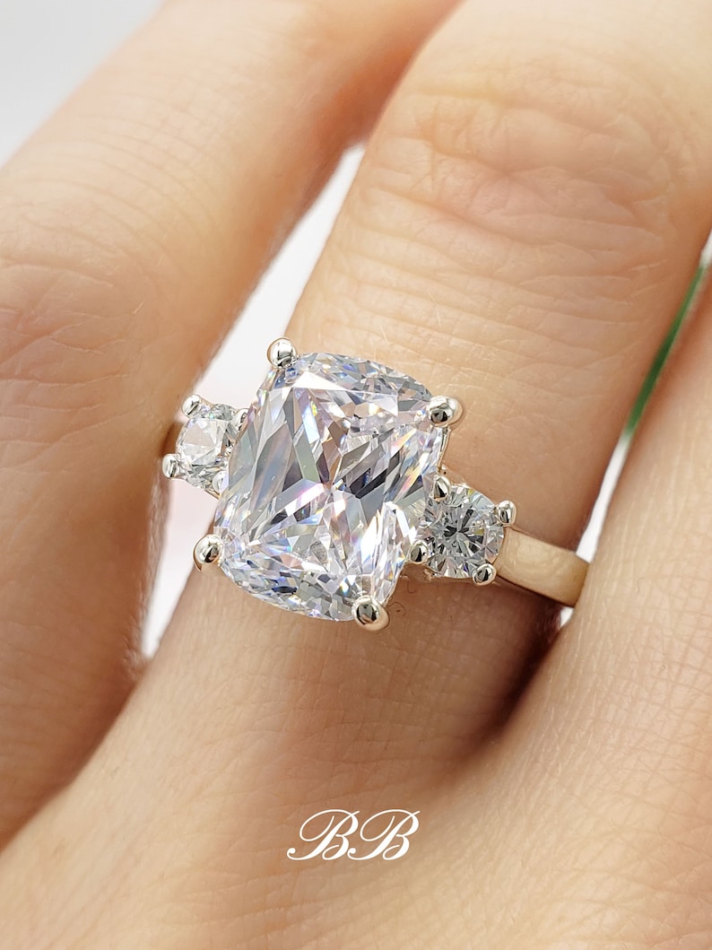 May include: A gold ring with a large, cushion-cut cubic zirconia center stone and two smaller round cubic zirconia accent stones.