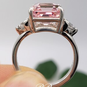 Puede incluir: Un anillo plateado con una gran gema rectangular de color rosa p&aacute;lido en el centro, flanqueada por dos gemas m&aacute;s peque&ntilde;as y transparentes. El anillo se sostiene, mostrando la banda y el engaste de las piedras. Las iniciales "BB" son visibles.