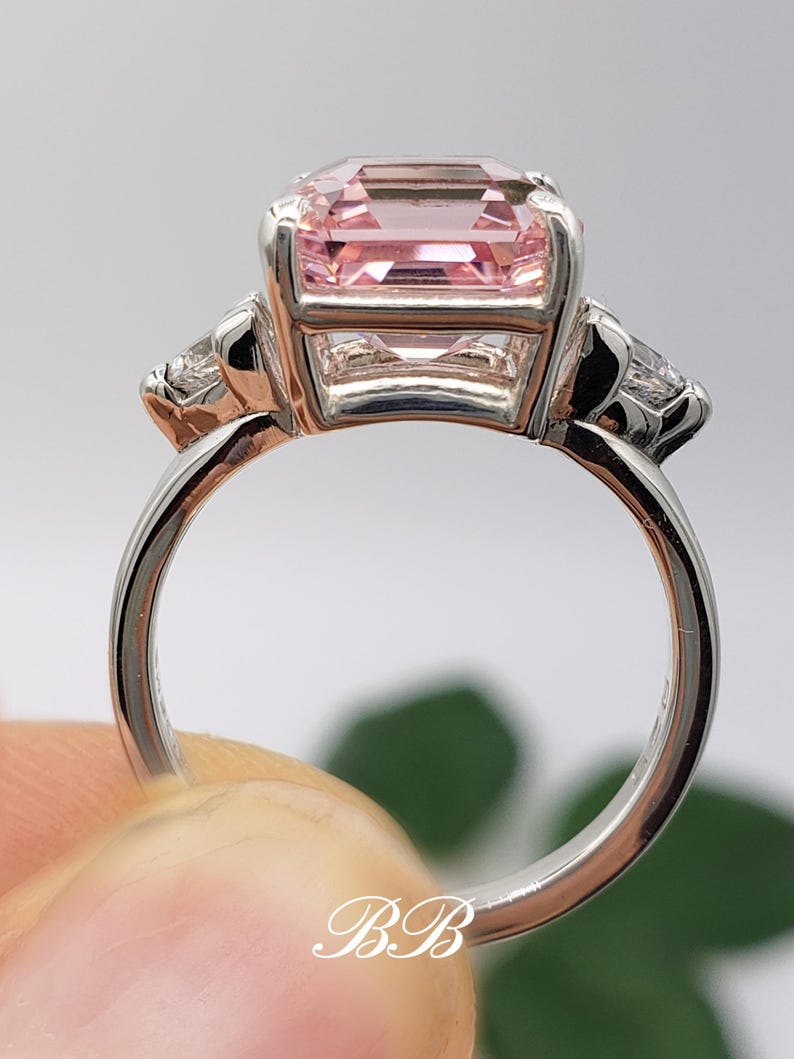 Puede incluir: Un anillo plateado con una gran gema rectangular de color rosa p&aacute;lido en el centro, flanqueada por dos gemas m&aacute;s peque&ntilde;as y transparentes. El anillo se sostiene, mostrando la banda y el engaste de las piedras. Las iniciales "BB" son visibles.