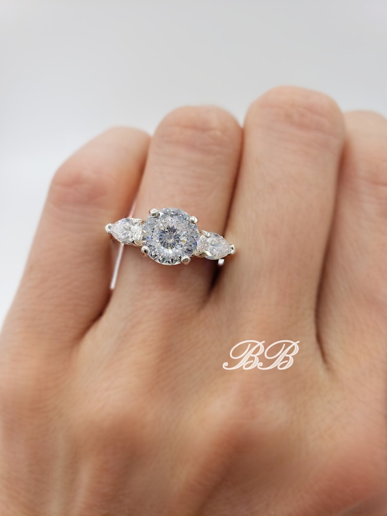 May include: A silver ring with a large round center stone and two pear-shaped stones on either side.