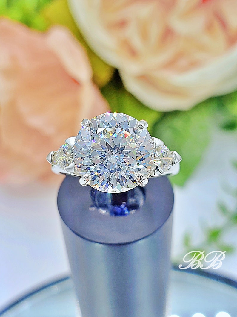 May include: A dazzling engagement ring featuring a large, round-cut diamond as the centerpiece, flanked by smaller, triangular accent stones. The ring is set in a silver band, showcasing the brilliance of the diamonds. The ring is displayed on a dark blue stand.
