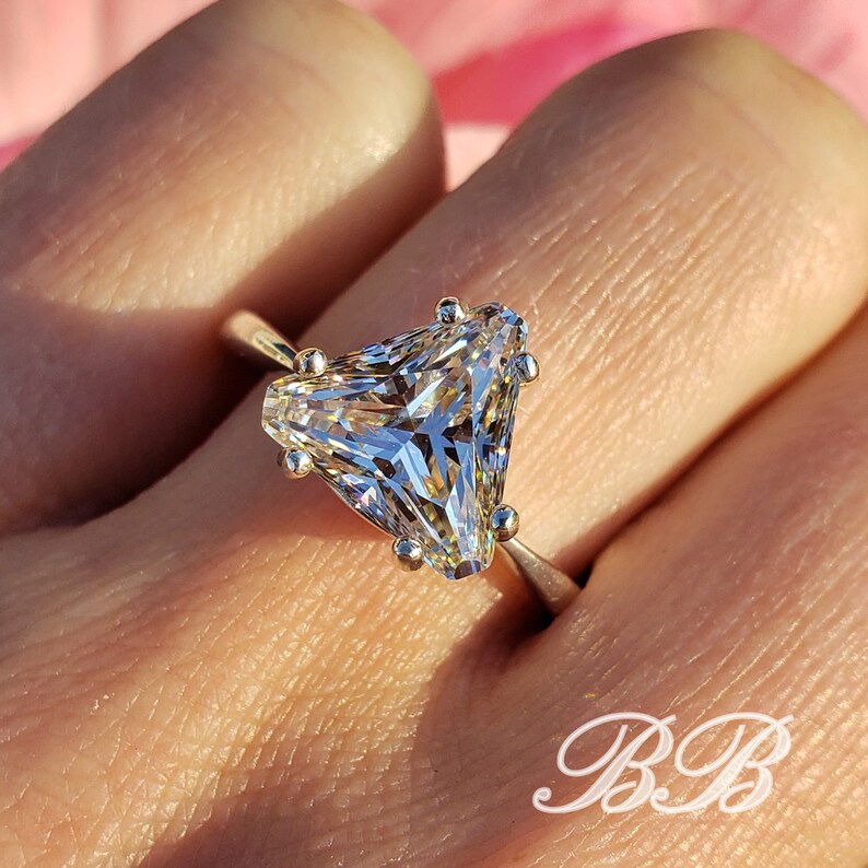 May include: A gold ring featuring a large, clear, triangular-cut gemstone. The gemstone is held in place by six small prongs. The ring is set against a soft, blurred background.