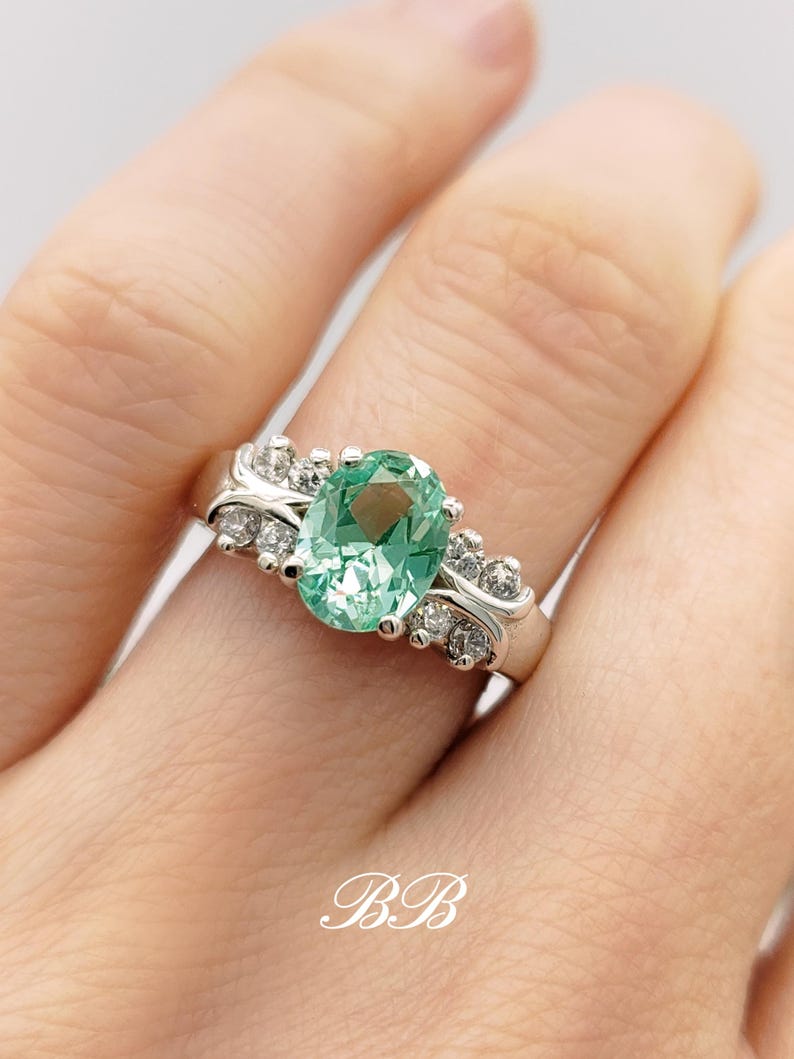 May include: A silver ring with a large oval green gemstone surrounded by smaller clear gemstones. The ring is set on a white background.