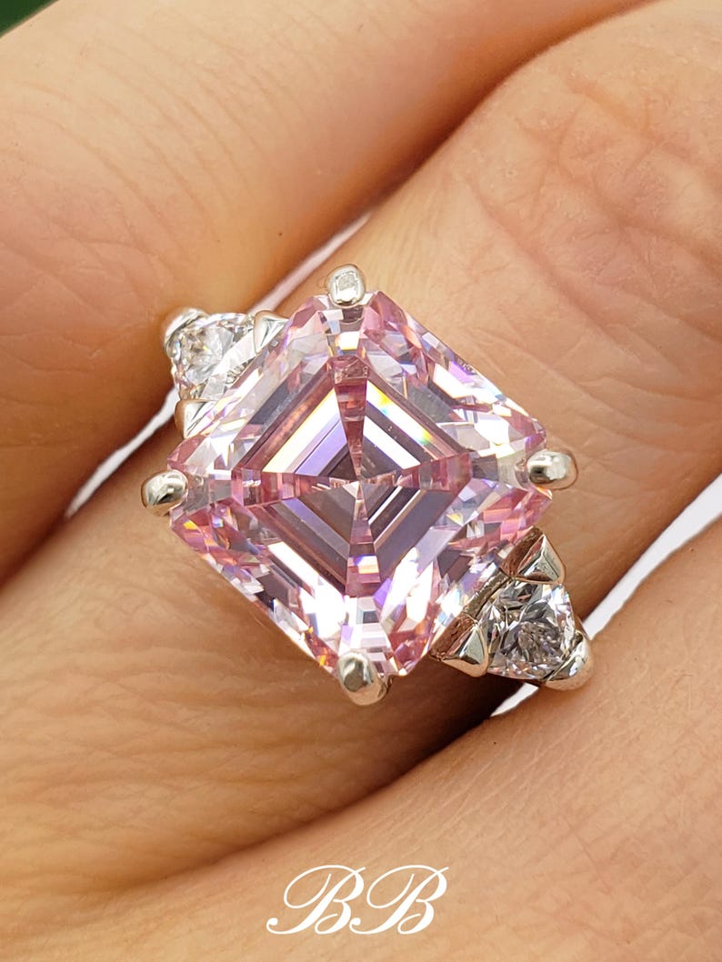 Puede incluir: Un anillo de plata con una gran gema rosa de talla cuadrada como pieza central. Dos gemas blancas m&aacute;s peque&ntilde;as flanquean la piedra principal. El anillo tiene un dise&ntilde;o delicado. Las iniciales "BB" son visibles.