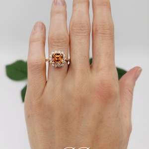 Pode incluir: Um anel de ouro com uma pedra preciosa laranja quadrada cercada por pedras preciosas menores e transparentes.
