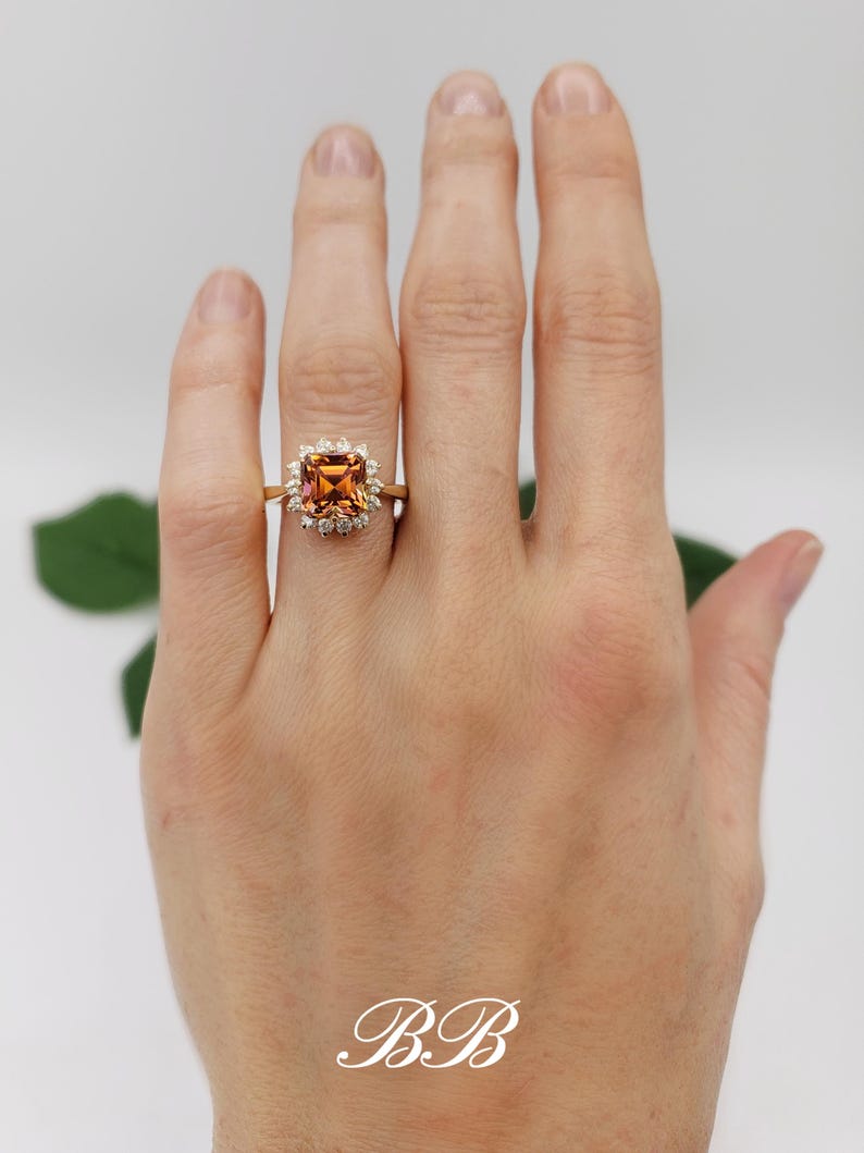 Pode incluir: Um anel de ouro com uma pedra preciosa laranja quadrada cercada por pedras preciosas menores e transparentes.