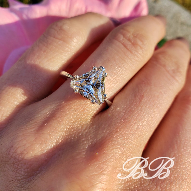 May include: A silver ring featuring a large, triangular-cut, clear gemstone. The ring's band is thin and delicate, with the gemstone held in place by multiple prongs. The ring is shown on a hand, with a soft pink background.