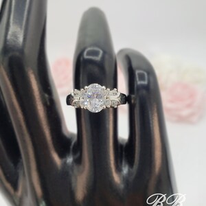 May include: A silver ring with a large oval-shaped clear stone surrounded by smaller clear stones. The ring is on a black ring display stand.