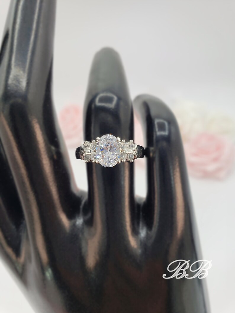 May include: A silver ring with a large oval-shaped clear stone surrounded by smaller clear stones. The ring is on a black ring display stand.