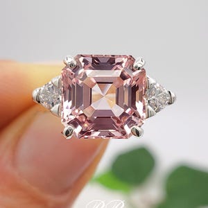 Puede incluir: Primer plano de un anillo plateado con una gran gema rosa de corte cuadrado y dos gemas blancas triangulares m&aacute;s peque&ntilde;as. La gema rosa es el punto focal, con una apariencia clara y facetada. El dise&ntilde;o es elegante y refinado.