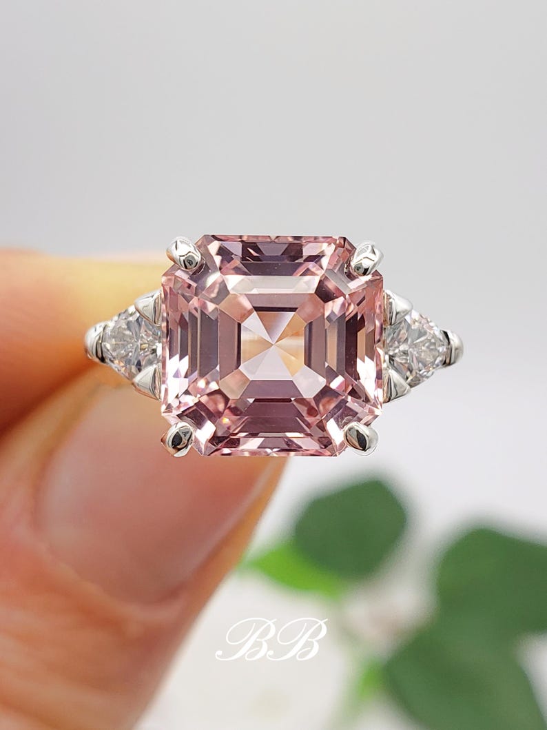 Puede incluir: Primer plano de un anillo plateado con una gran gema rosa de corte cuadrado y dos gemas blancas triangulares m&aacute;s peque&ntilde;as. La gema rosa es el punto focal, con una apariencia clara y facetada. El dise&ntilde;o es elegante y refinado.
