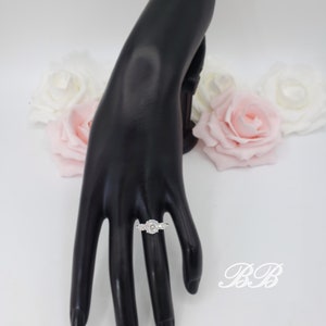 May include: A silver ring with a large oval-shaped clear stone surrounded by smaller clear stones. The ring is on a black hand model.
