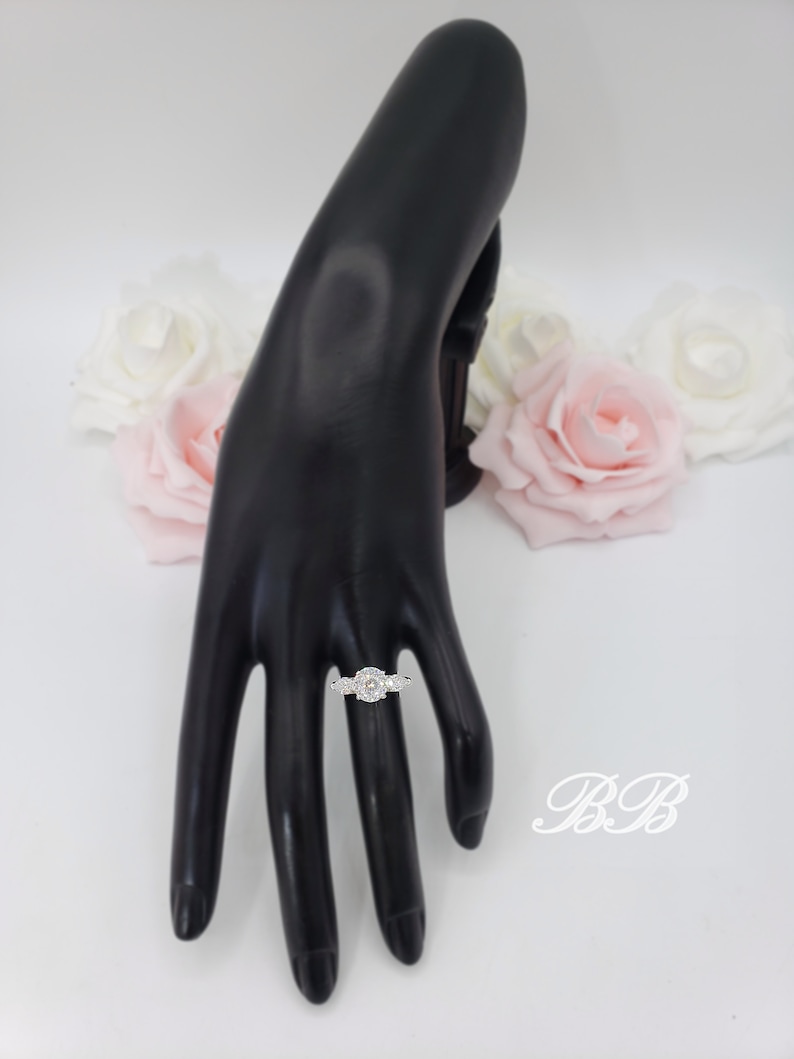 May include: A silver ring with a large oval-shaped clear stone surrounded by smaller clear stones. The ring is on a black hand model.
