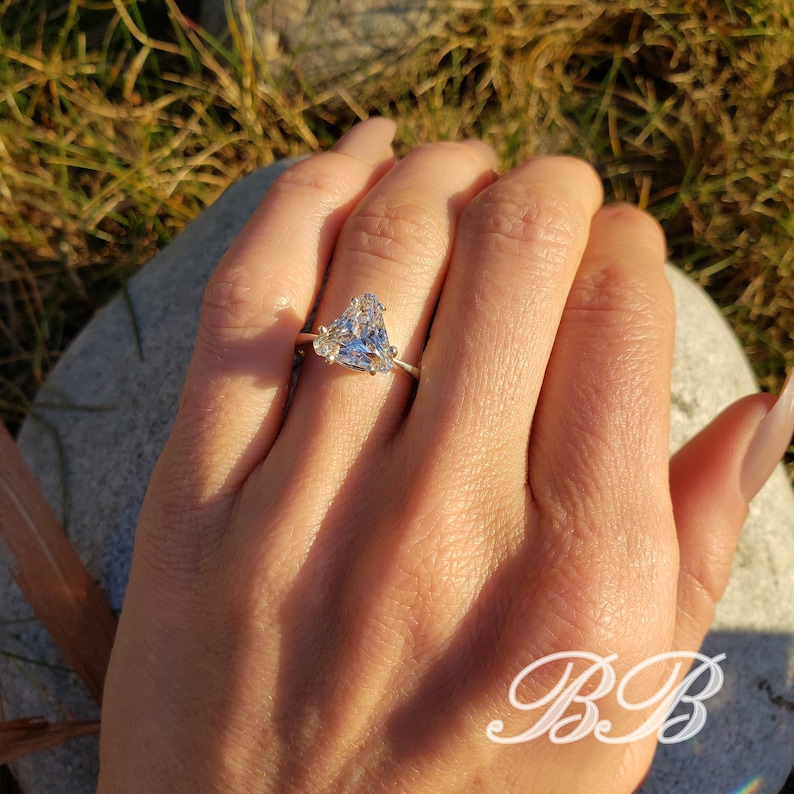 May include: A silver ring featuring a large, triangular-cut diamond as the primary stone, with smaller accent stones on either side. The ring is set on a slender band. The image is taken in natural light, highlighting the sparkle of the diamonds.
