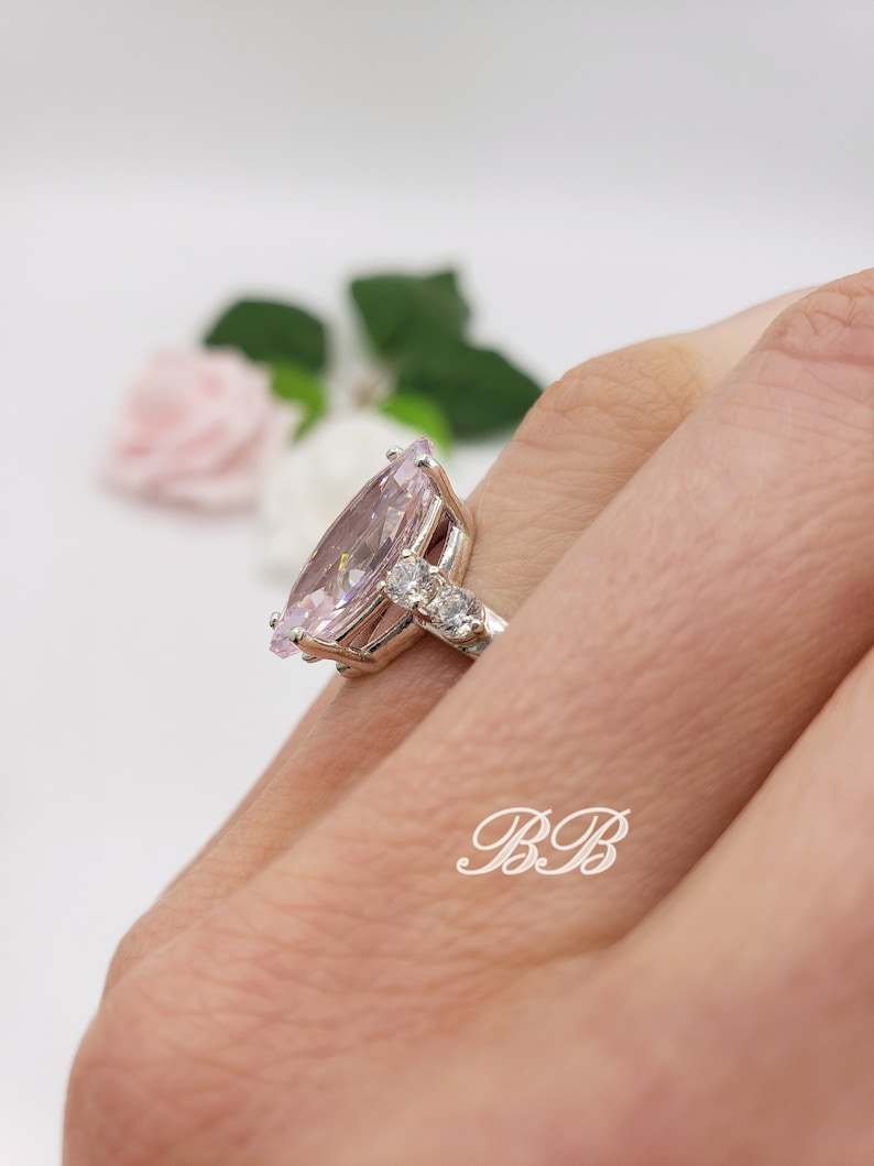 K&ouml;nnte beinhalten: Ein silberner Ring mit einem gro&szlig;en birnenf&ouml;rmigen rosafarbenen Edelstein und kleineren klaren Edelsteinen auf der Ringschiene.