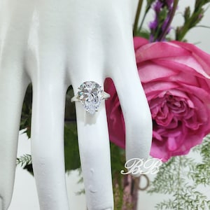 May include: A pear-shaped diamond engagement ring with a silver band and two smaller diamonds on the side. The ring is on a white mannequin hand.