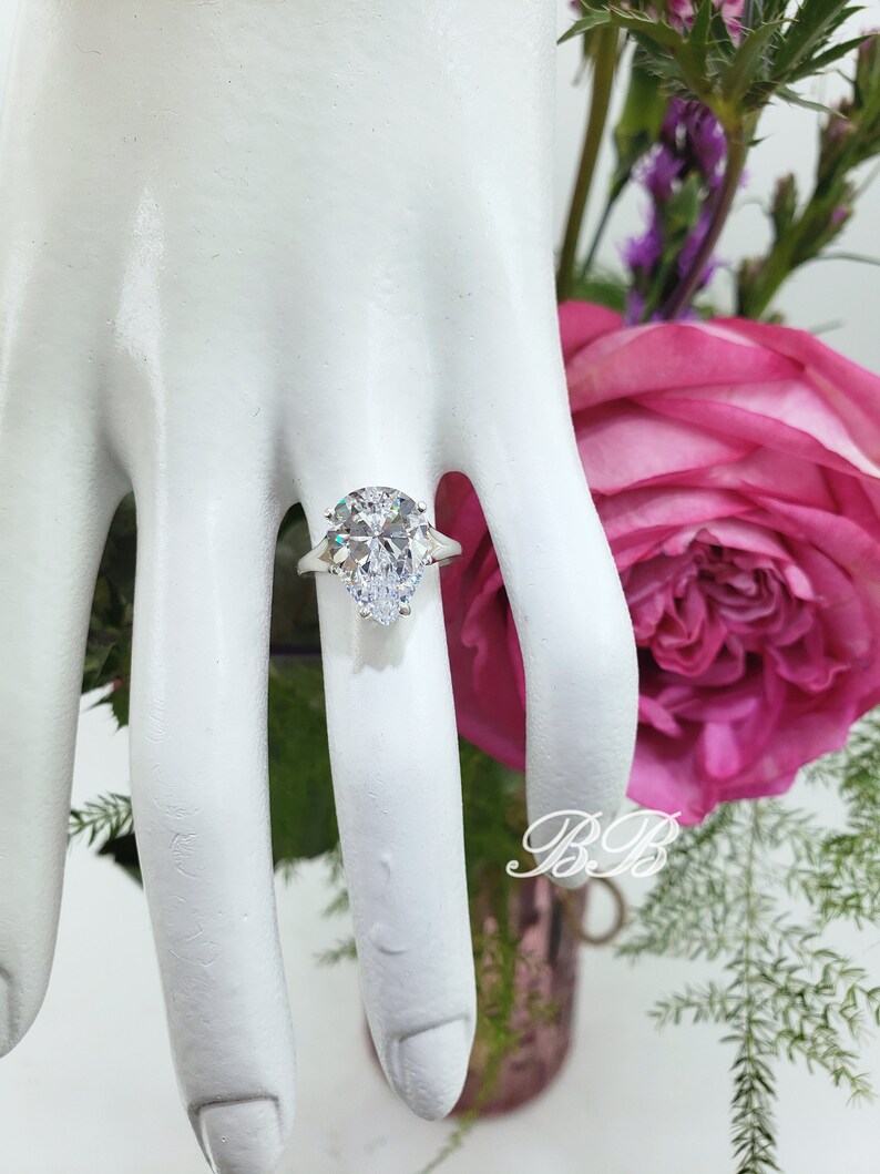 May include: A pear-shaped diamond engagement ring with a silver band and two smaller diamonds on the side. The ring is on a white mannequin hand.