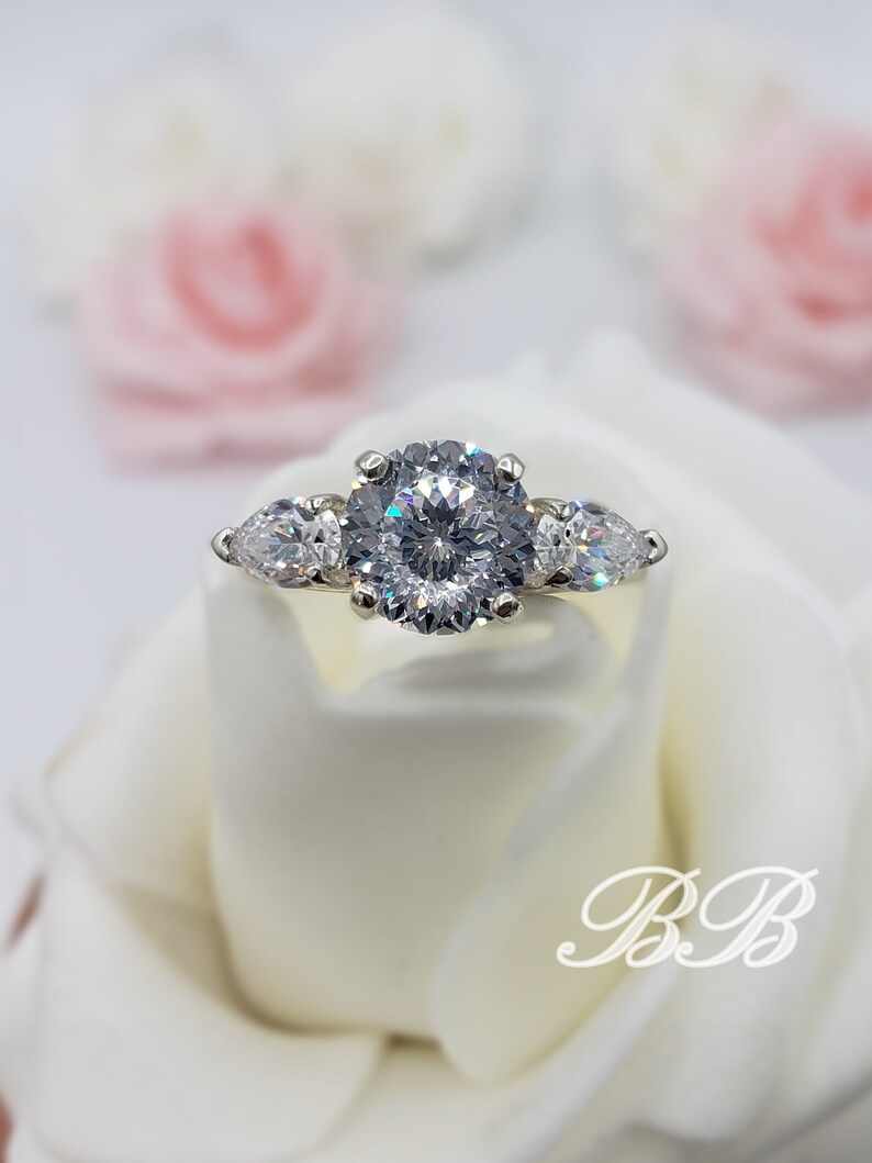 May include: A silver ring with a large round center stone and two pear-shaped stones on either side. The ring is set on a white background.