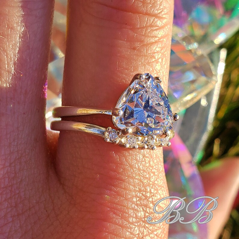 May include: A silver ring set with a large, triangular, clear gemstone surrounded by smaller clear gemstones. The ring is worn on a finger.