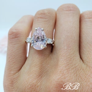 Puede incluir: Un anillo de compromiso de forma de pera con una gran piedra central rosa y dos piedras redondas m&aacute;s peque&ntilde;as a cada lado. El anillo est&aacute; engastado en plata.