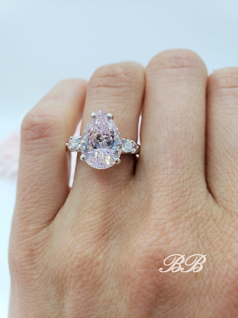 Puede incluir: Un anillo de compromiso de forma de pera con una gran piedra central rosa y dos piedras redondas m&aacute;s peque&ntilde;as a cada lado. El anillo est&aacute; engastado en plata.