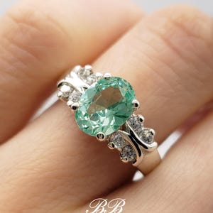 Puede incluir: Un anillo de plata con una gran piedra preciosa ovalada verde rodeada de peque&ntilde;as piedras preciosas transparentes. El anillo se lleva en un dedo.