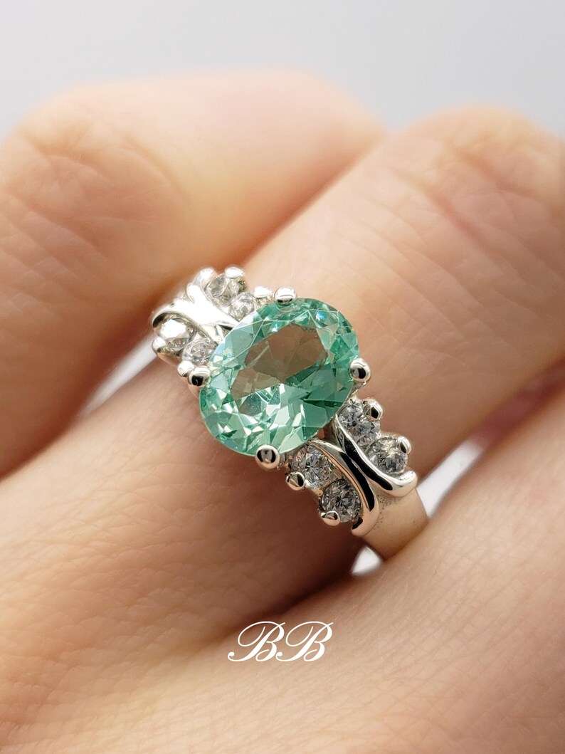 Puede incluir: Un anillo de plata con una gran piedra preciosa ovalada verde rodeada de peque&ntilde;as piedras preciosas transparentes. El anillo se lleva en un dedo.