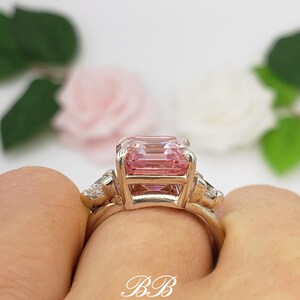 Puede incluir: Un anillo de oro con una gran gema rosa rectangular y piedras m&aacute;s peque&ntilde;as y claras. El anillo es sostenido por una mano, con las iniciales "BB" en la banda. El fondo presenta flores y follaje borrosos.