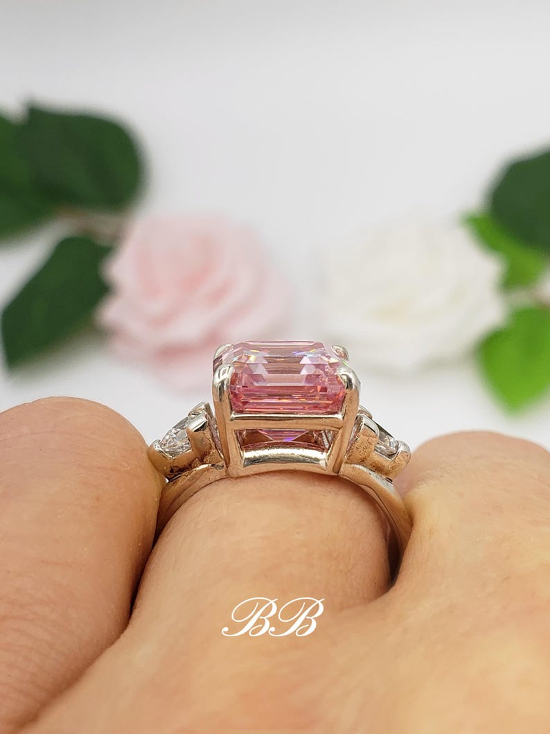Puede incluir: Un anillo de oro con una gran gema rosa rectangular y piedras m&aacute;s peque&ntilde;as y claras. El anillo es sostenido por una mano, con las iniciales "BB" en la banda. El fondo presenta flores y follaje borrosos.