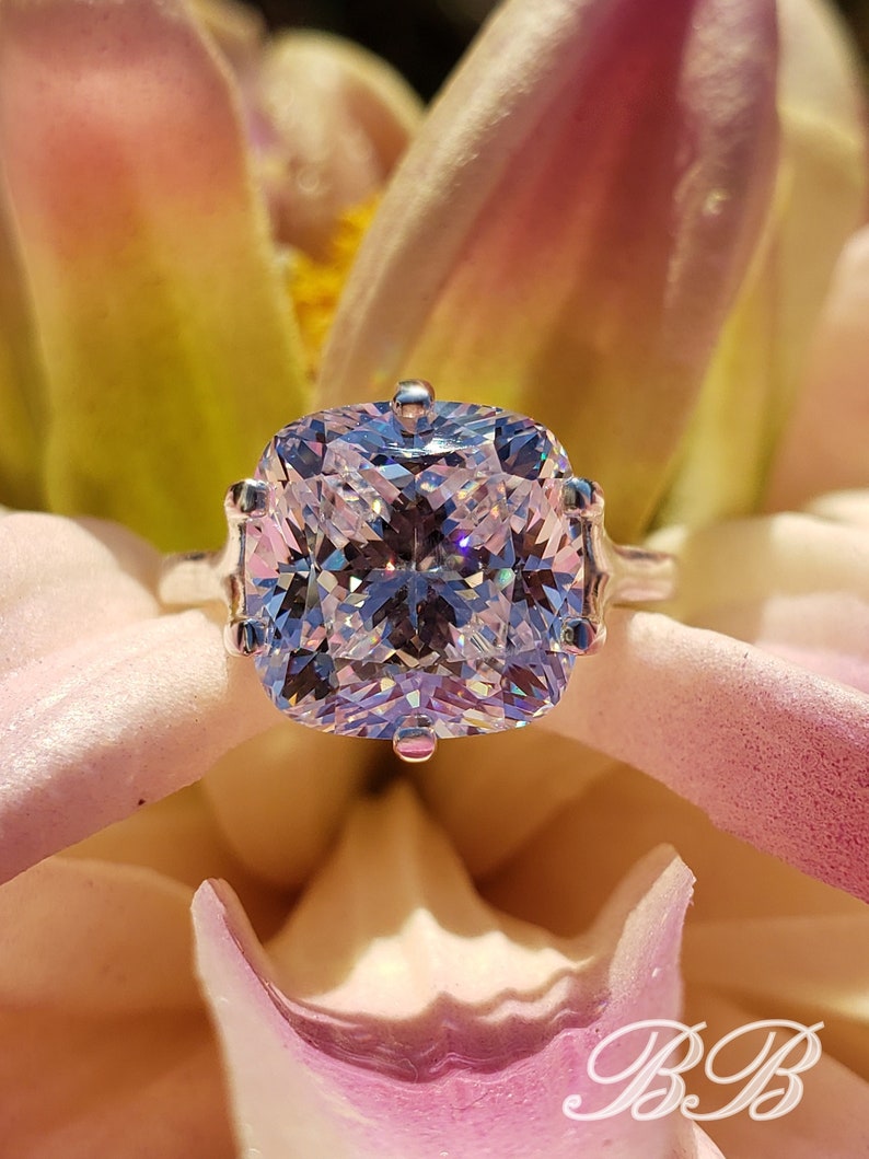 May include: A silver ring with a large, clear, cushion-cut gemstone. The ring is set against a pink and white flower.