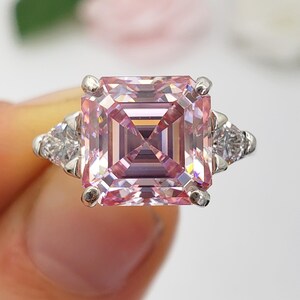 Puede incluir: Un anillo de plata con una gran gema rosa p&aacute;lido de forma cuadrada y talla escalonada. Dos gemas triangulares m&aacute;s peque&ntilde;as y transparentes flanquean la piedra principal. El anillo se presenta sobre un fondo suave y borroso.