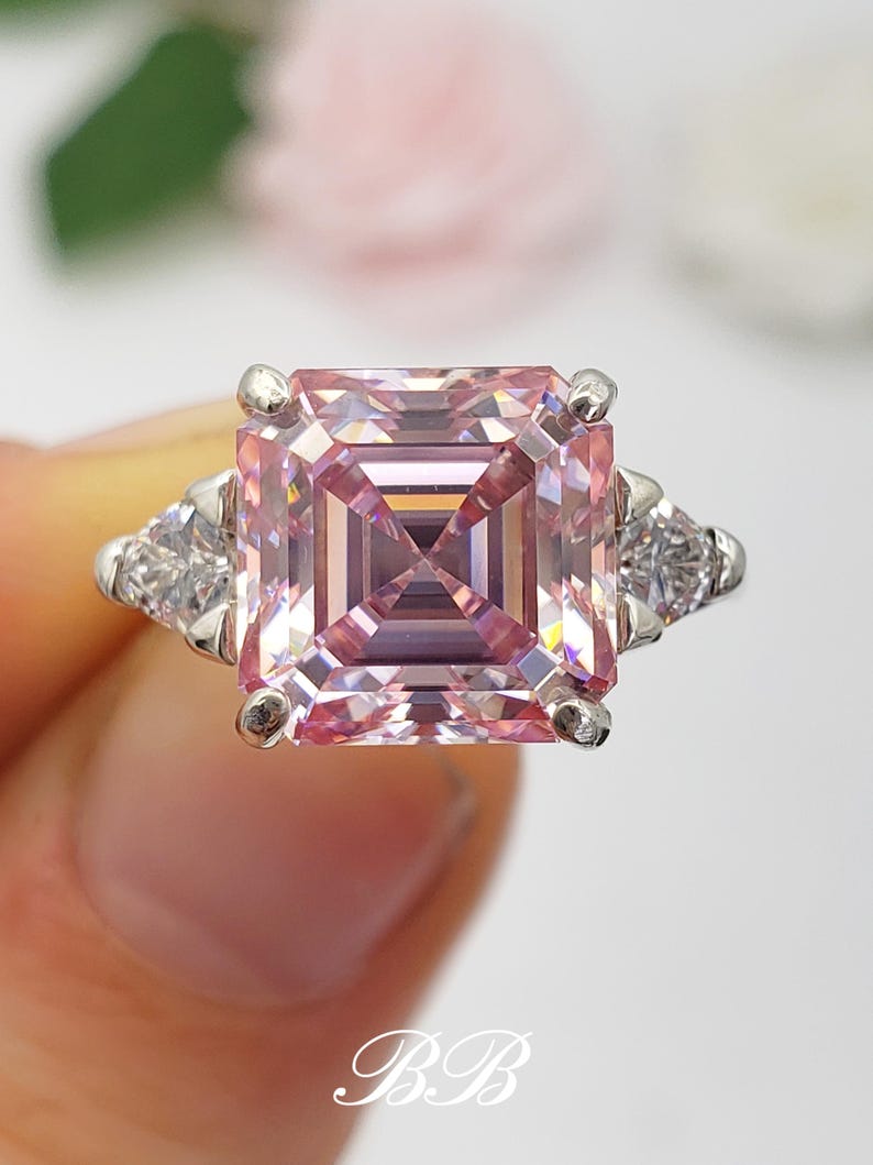 Puede incluir: Un anillo de plata con una gran gema rosa p&aacute;lido de forma cuadrada y talla escalonada. Dos gemas triangulares m&aacute;s peque&ntilde;as y transparentes flanquean la piedra principal. El anillo se presenta sobre un fondo suave y borroso.