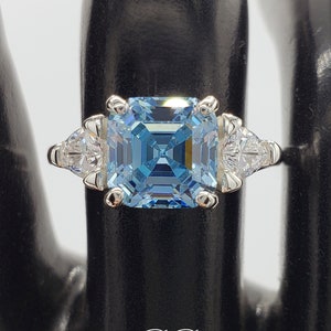 May include: A silver ring with a large, square, light blue gemstone surrounded by two smaller, triangular, clear gemstones.