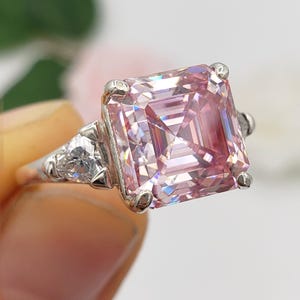 Puede incluir: Un anillo de plata con una gran gema rosa de corte cuadrado como pieza central. Dos piedras claras triangulares m&aacute;s peque&ntilde;as flanquean la piedra principal. El anillo se presenta sobre un fondo suave y borroso.
