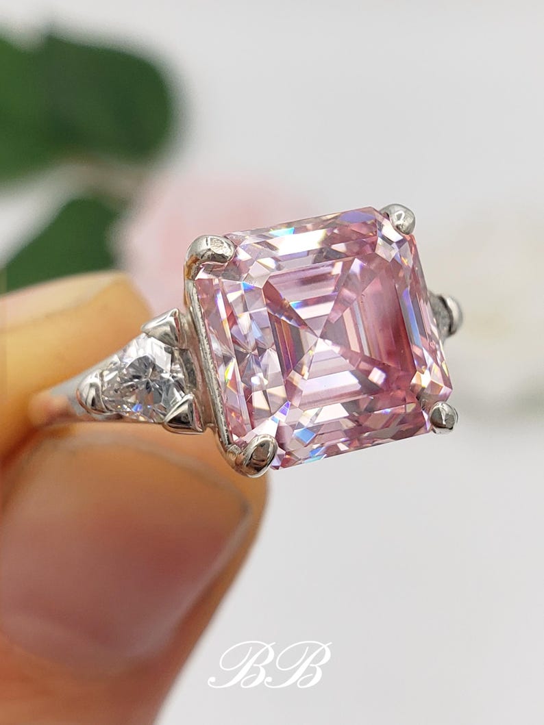 Puede incluir: Un anillo de plata con una gran gema rosa de corte cuadrado como pieza central. Dos piedras claras triangulares m&aacute;s peque&ntilde;as flanquean la piedra principal. El anillo se presenta sobre un fondo suave y borroso.