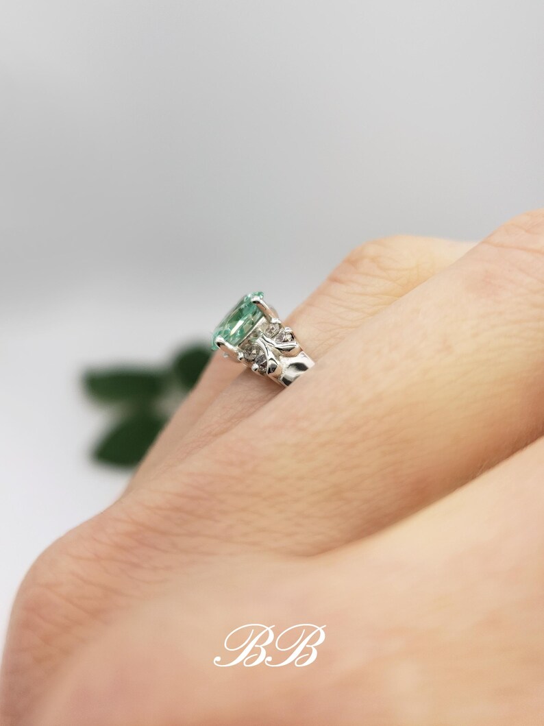 Puede incluir: Un anillo de plata con una piedra preciosa verde y detalles florales intrincados. El anillo est&aacute; en un dedo.