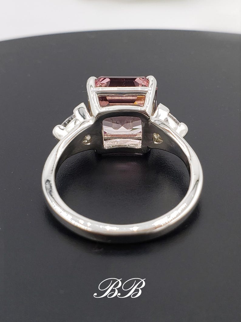 Puede incluir: Un anillo de plata con una gema rosa rectangular como elemento central. El anillo tiene gemas claras m&aacute;s peque&ntilde;as a ambos lados de la piedra principal. La banda pulida es lisa, con las iniciales "BB" en escritura en la base.